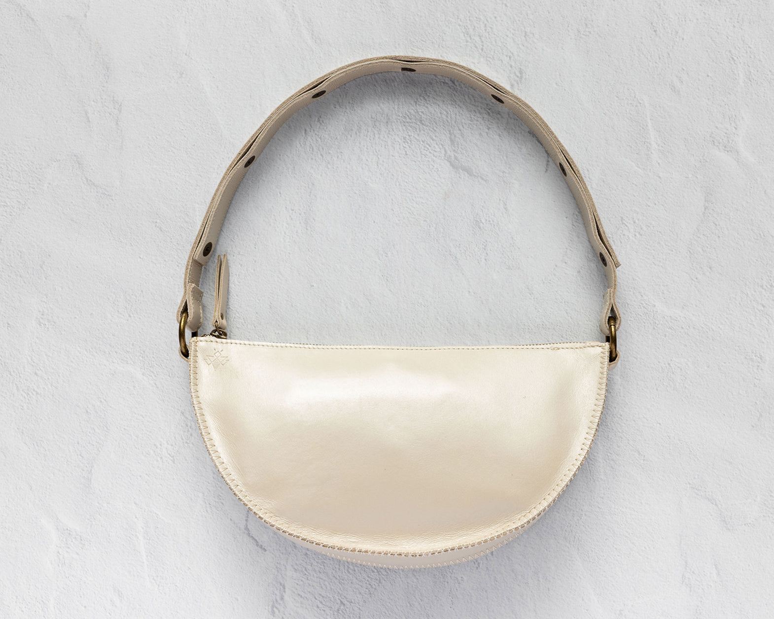 A cream-colored crescent-shaped shoulder bag with a glossy finish, featuring a matching strap with black polka dots and gold-tone hardware, set against a textured light gray background.