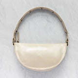 A cream-colored crescent-shaped shoulder bag with a glossy finish, featuring a matching strap with black polka dots and gold-tone hardware, set against a textured light gray background.
