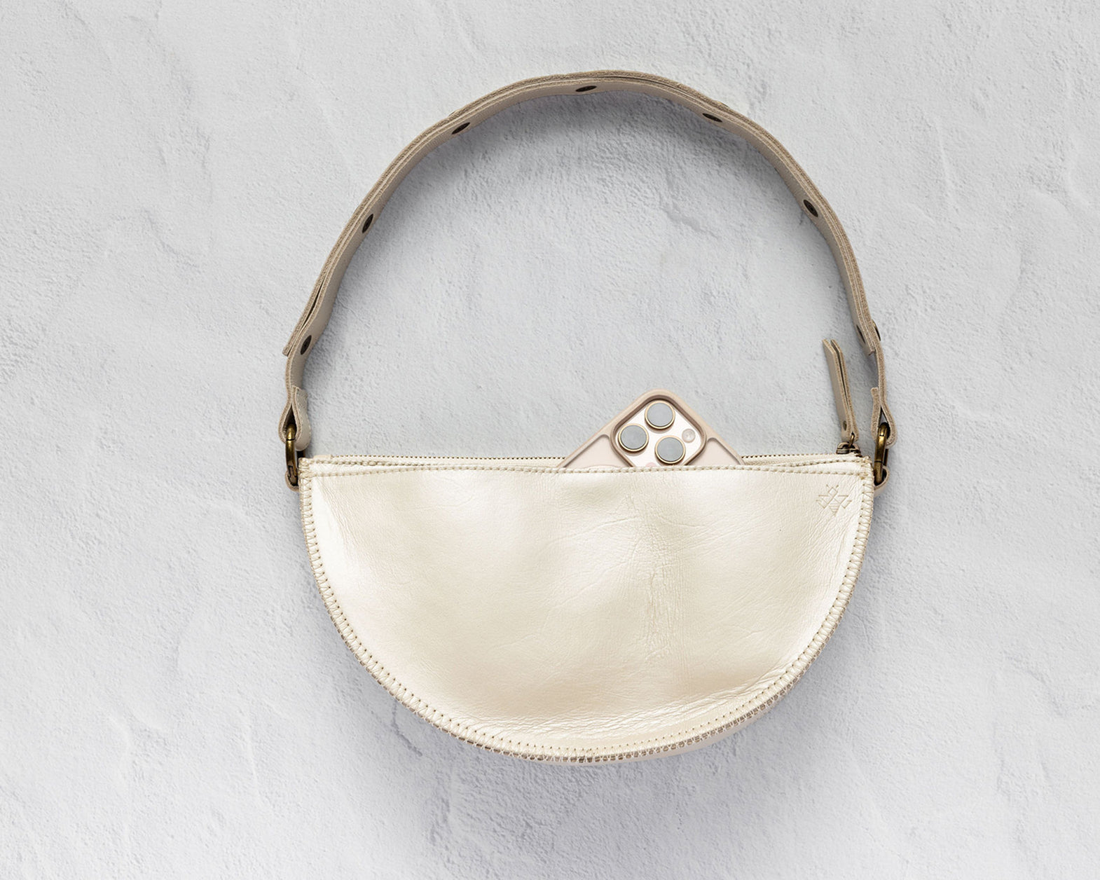 A cream-colored leather crescent-shaped shoulder bag with a matching leather strap and gold-tone hardware, shown against a textured light gray background.