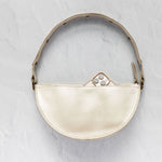 A cream-colored leather crescent-shaped shoulder bag with a matching leather strap and gold-tone hardware, shown against a textured light gray background.