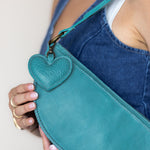 A close-up of a person wearing a blue denim apron, holding a teal leather crossbody bag with a textured heart-shaped charm.