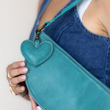 A close-up of a person wearing a blue denim apron, holding a teal leather crossbody bag with a textured heart-shaped charm.