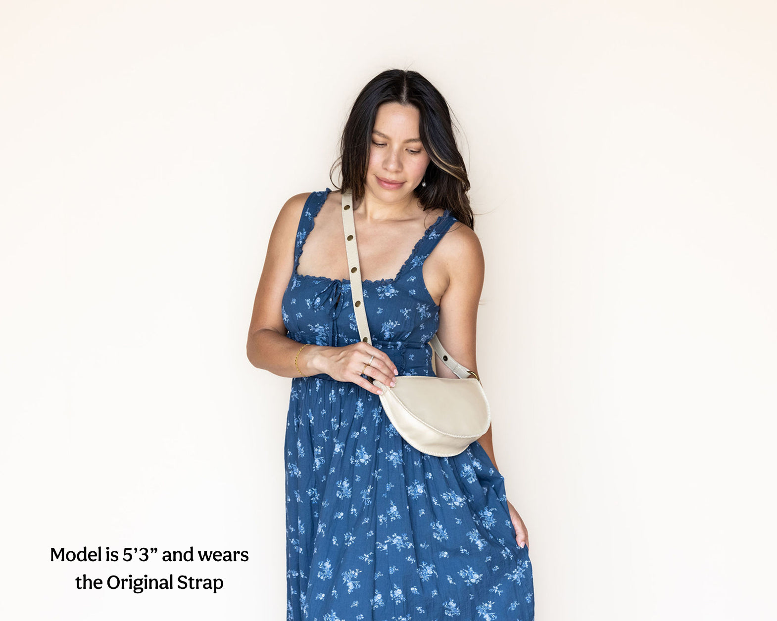 A woman wearing a blue floral dress and a cream crescent-shaped shoulder bag with a long strap, standing against a plain off-white wall. Text in the lower-left corner reads, 'Model is 5'3" and wears the Original Strap'.