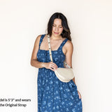 A woman wearing a blue floral dress and a cream crescent-shaped shoulder bag with a long strap, standing against a plain off-white wall. Text in the lower-left corner reads, 'Model is 5'3" and wears the Original Strap'.