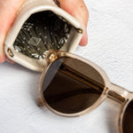 A close-up of a hand holding a small beige leather pouch with a patterned lining, next to a pair of round sunglasses with dark lenses and a gold-tone frame, on a textured white surface.