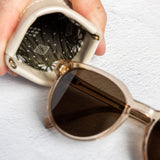 A close-up of a hand holding a small beige leather pouch with a patterned lining, next to a pair of round sunglasses with dark lenses and a gold-tone frame, on a textured white surface.