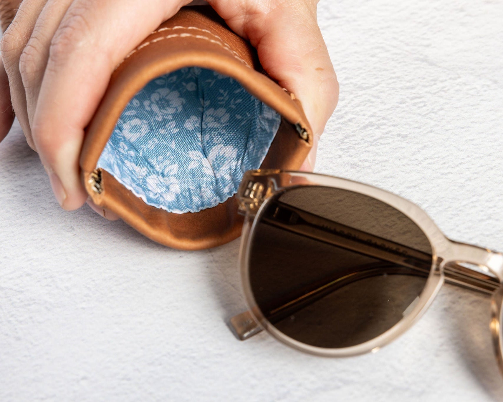 A close-up of a hand holding an open brown leather sunglasses case with a blue floral lining, with a pair of sunglasses resting beside it on a white textured surface.