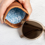 A close-up of a hand holding an open brown leather sunglasses case with a blue floral lining, with a pair of sunglasses resting beside it on a white textured surface.