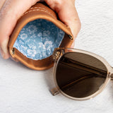 A close-up of a hand holding an open brown leather sunglasses case with a blue floral lining, with a pair of sunglasses resting beside it on a white textured surface.