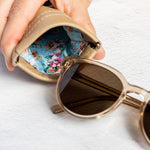 A close-up of a hand holding an open beige leather sunglasses case, showing a colorful floral-patterned interior, with a pair of round sunglasses resting inside.