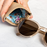 A close-up of a hand holding an open beige leather sunglasses case, showing a colorful floral-patterned interior, with a pair of round sunglasses resting inside.