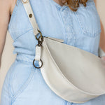 A light beige faux-leather crescent-shaped shoulder bag with a matching strap and dark metal hardware, shown worn cross-body over a light blue denim dress.