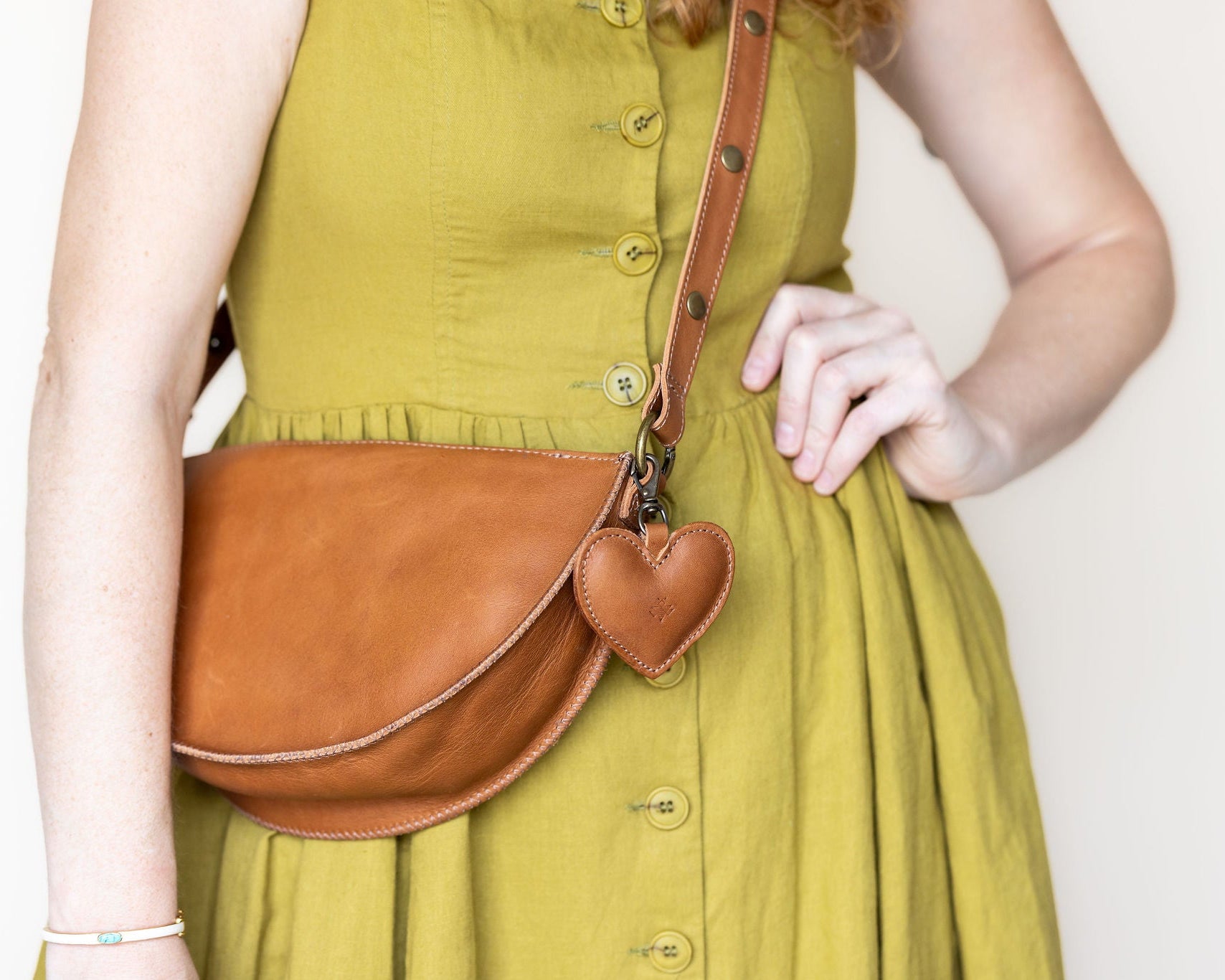 A person wearing a mustard yellow dress carries a brown leather half-moon crossbody bag with a small heart-shaped charm on a plain light background.