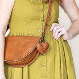A person wearing a mustard yellow dress carries a brown leather half-moon crossbody bag with a small heart-shaped charm on a plain light background.