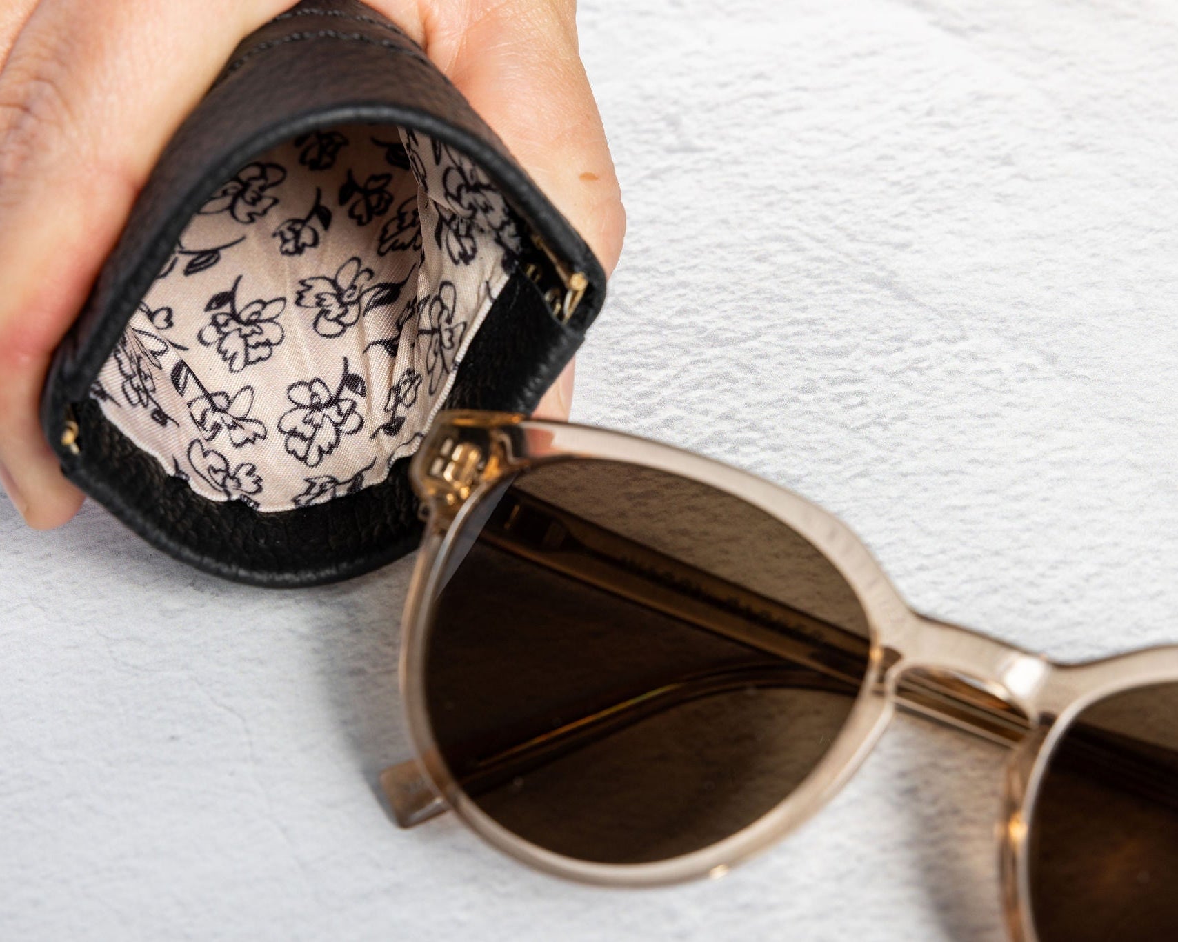 A close-up of a hand holding an open black sunglasses case with a beige floral lining next to a pair of round sunglasses with gold frames and dark lenses on a textured white background.