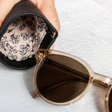 A close-up of a hand holding an open black sunglasses case with a beige floral lining next to a pair of round sunglasses with gold frames and dark lenses on a textured white background.