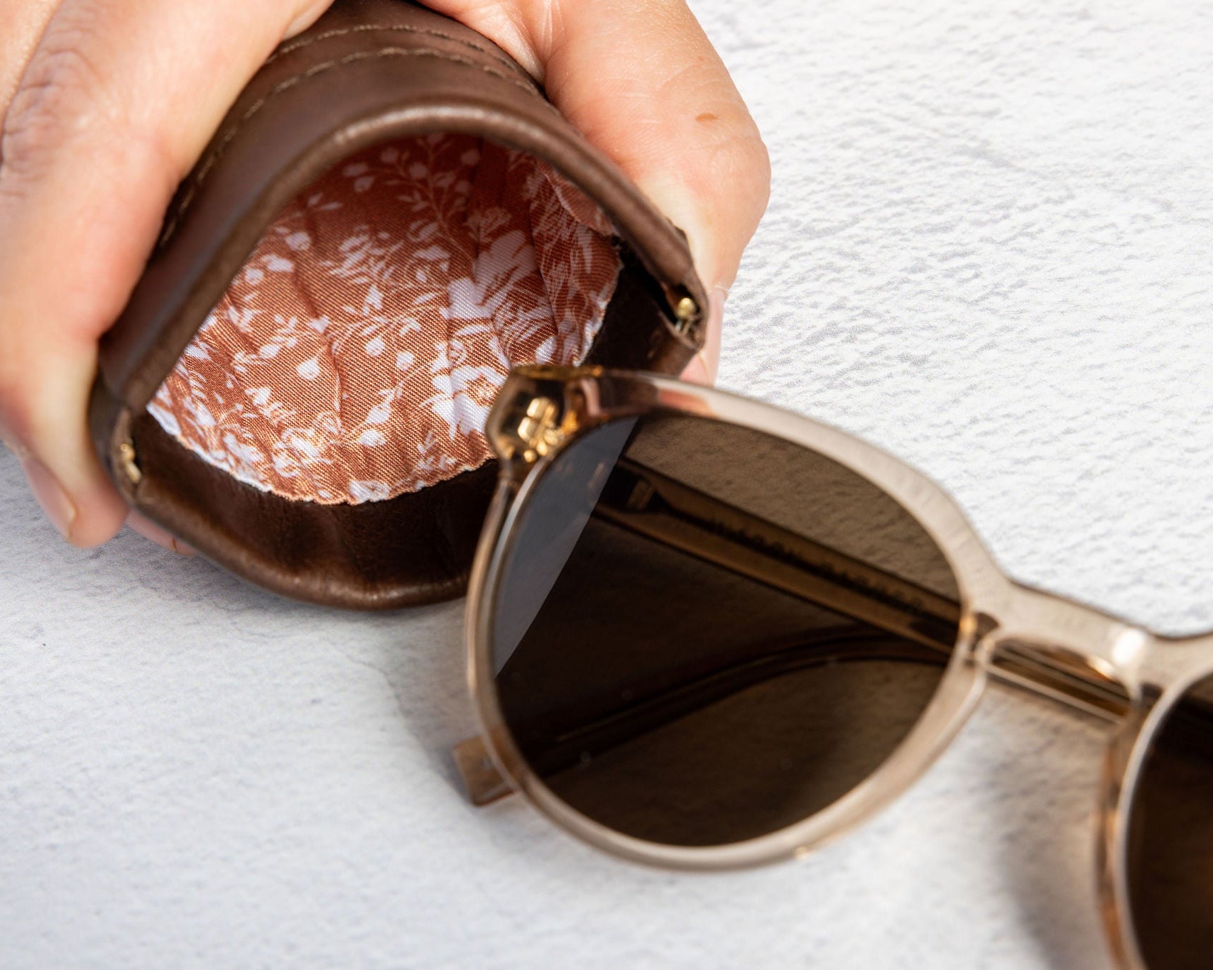 A close-up of a hand holding an open brown leather sunglasses case next to a pair of round sunglasses with dark lenses and a gold-tone frame, set against a textured white background.