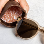 A close-up of a hand holding an open brown leather sunglasses case next to a pair of round sunglasses with dark lenses and a gold-tone frame, set against a textured white background.