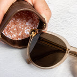A close-up of a hand holding an open brown leather sunglasses case next to a pair of round sunglasses with dark lenses and a gold-tone frame, set against a textured white background.