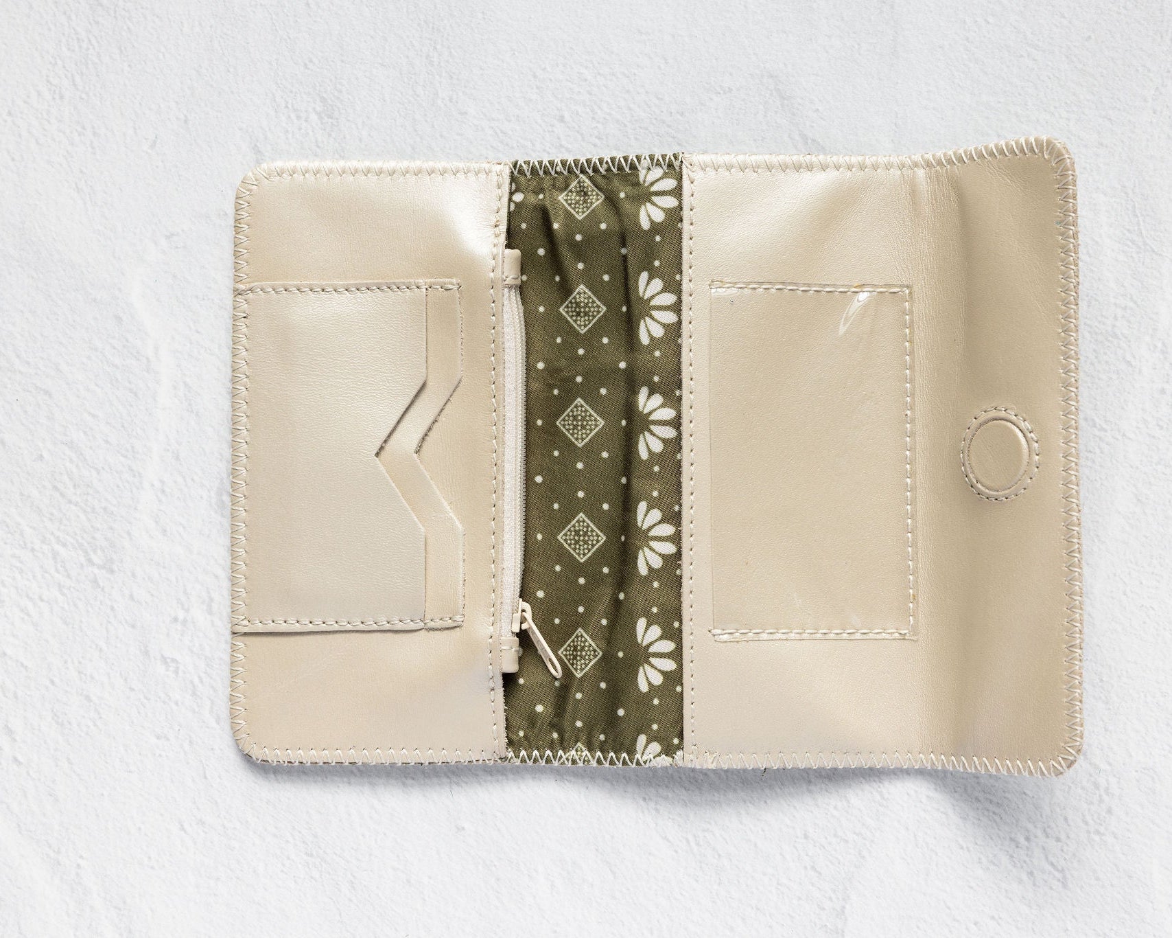 An open cream-colored leather wallet with a central olive green patterned fabric divider, displayed on a textured white background.