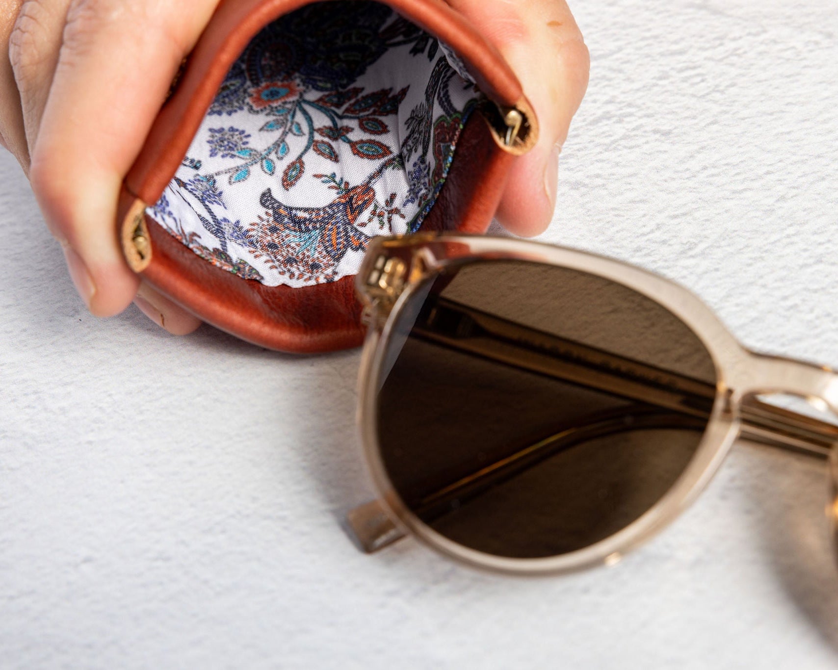 A close-up of a hand holding an open brown leather sunglasses case with a colorful paisley-patterned lining, with a pair of sunglasses resting beside it on a textured white surface.