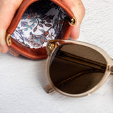 A close-up of a hand holding an open brown leather sunglasses case with a colorful paisley-patterned lining, with a pair of sunglasses resting beside it on a textured white surface.