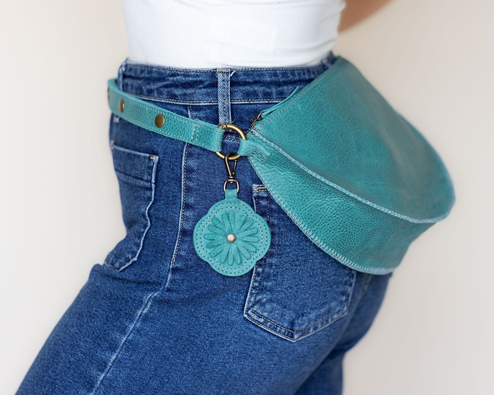 A teal leather belt bag worn around blue jeans, featuring a matching fabric flower-shaped charm with a small gold-tone ring and snap closure.
