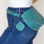 A teal leather belt bag worn around blue jeans, featuring a matching fabric flower-shaped charm with a small gold-tone ring and snap closure.