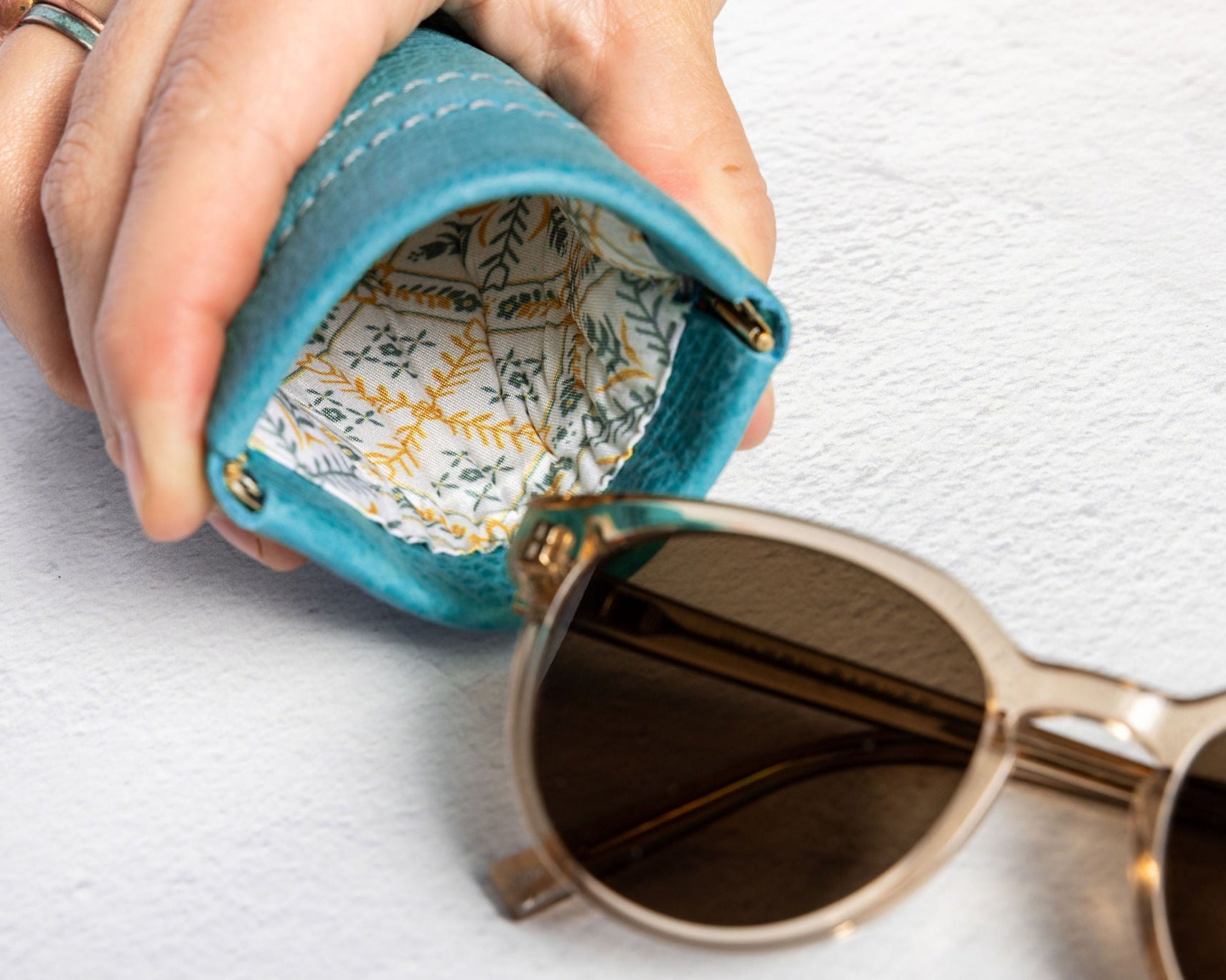 A close-up of a hand holding an open turquoise blue leather sunglasses case, with patterned lining visible inside and a pair of sunglasses resting outside on a white textured surface.