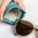 A close-up of a hand holding an open turquoise blue leather sunglasses case, with patterned lining visible inside and a pair of sunglasses resting outside on a white textured surface.