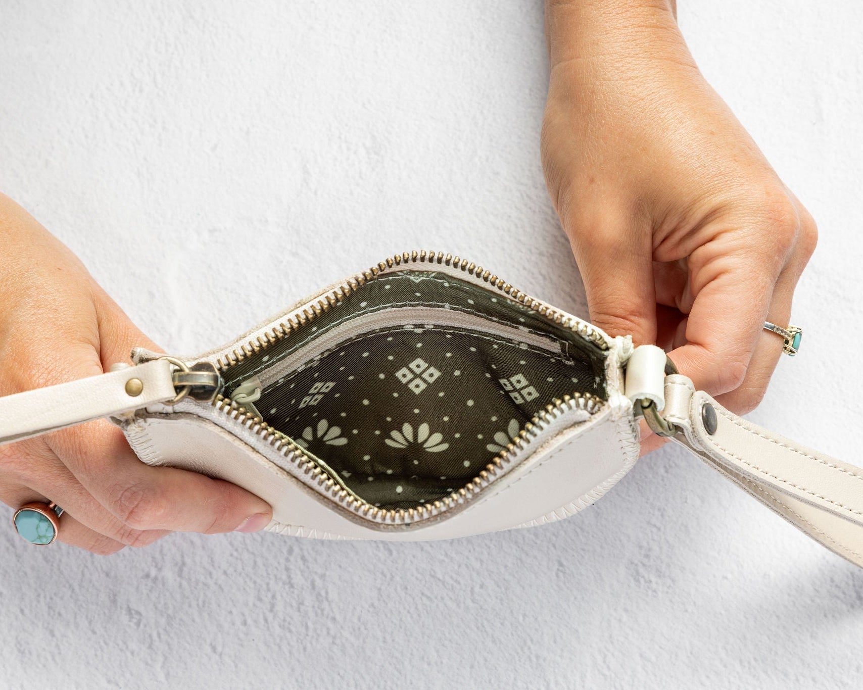 A close-up of a person's hands holding open a small white leather wristlet pouch, showing a dark patterned interior lining with a white geometric print.