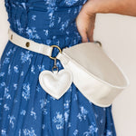 A cream-colored faux leather belt bag with a heart-shaped charm, worn around the waist of a person in a blue floral dress.