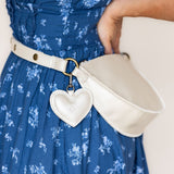 A cream-colored faux leather belt bag with a heart-shaped charm, worn around the waist of a person in a blue floral dress.