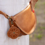 A tan leather fanny pack worn around a person's waist, featuring a decorative circular leather charm with a floral cutout pattern and a metal clasp. The person is wearing a white button-down shirt, with a blurred outdoor background.