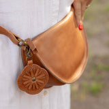 A tan leather fanny pack worn around a person's waist, featuring a decorative circular leather charm with a floral cutout pattern and a metal clasp. The person is wearing a white button-down shirt, with a blurred outdoor background.