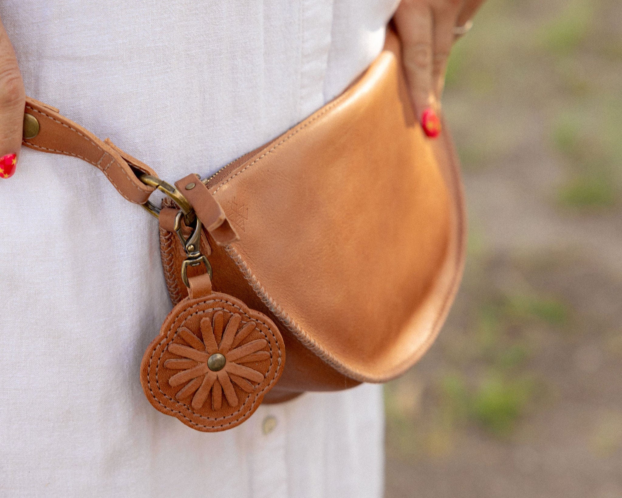 A tan leather fanny pack worn around a person's waist, featuring a decorative circular leather charm with a floral cutout pattern and a metal clasp. The person is wearing a white button-down shirt, with a blurred outdoor background.