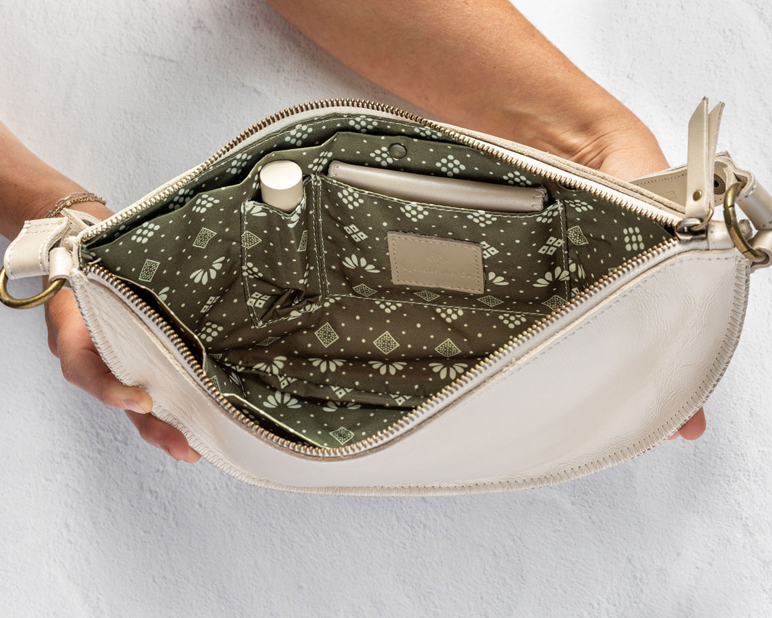 An open cream-colored leather clutch bag held by two hands, showcasing its olive green patterned interior lining with pockets and a zipper closure.