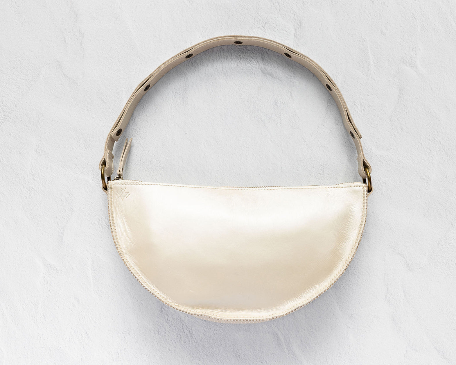 A cream-colored crescent-shaped shoulder bag with a glossy finish, featuring a matching strap with black polka dots and gold-tone hardware, set against a textured light gray background.