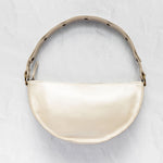 A cream-colored crescent-shaped shoulder bag with a glossy finish, featuring a matching strap with black polka dots and gold-tone hardware, set against a textured light gray background.