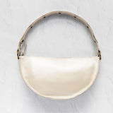 A cream-colored crescent-shaped shoulder bag with a glossy finish, featuring a matching strap with black polka dots and gold-tone hardware, set against a textured light gray background.