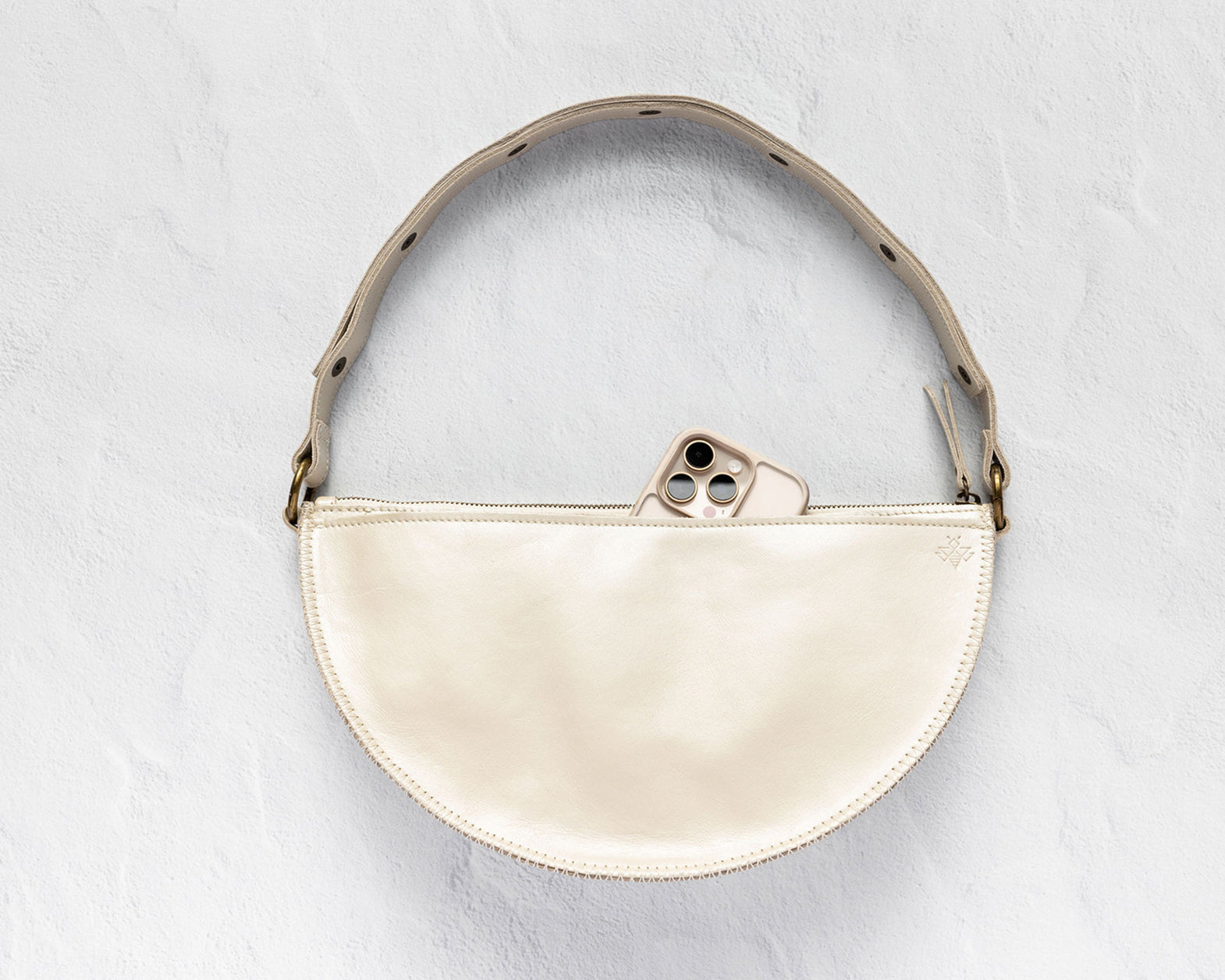 A cream-colored leather half-moon shoulder bag with a matching leather strap and gold-tone hardware, shown on a textured light gray background with a phone partially tucked into the zipper.