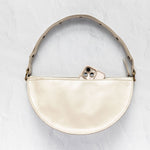A cream-colored leather half-moon shoulder bag with a matching leather strap and gold-tone hardware, shown on a textured light gray background with a phone partially tucked into the zipper.