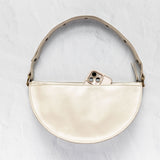 A cream-colored leather half-moon shoulder bag with a matching leather strap and gold-tone hardware, shown on a textured light gray background with a phone partially tucked into the zipper.