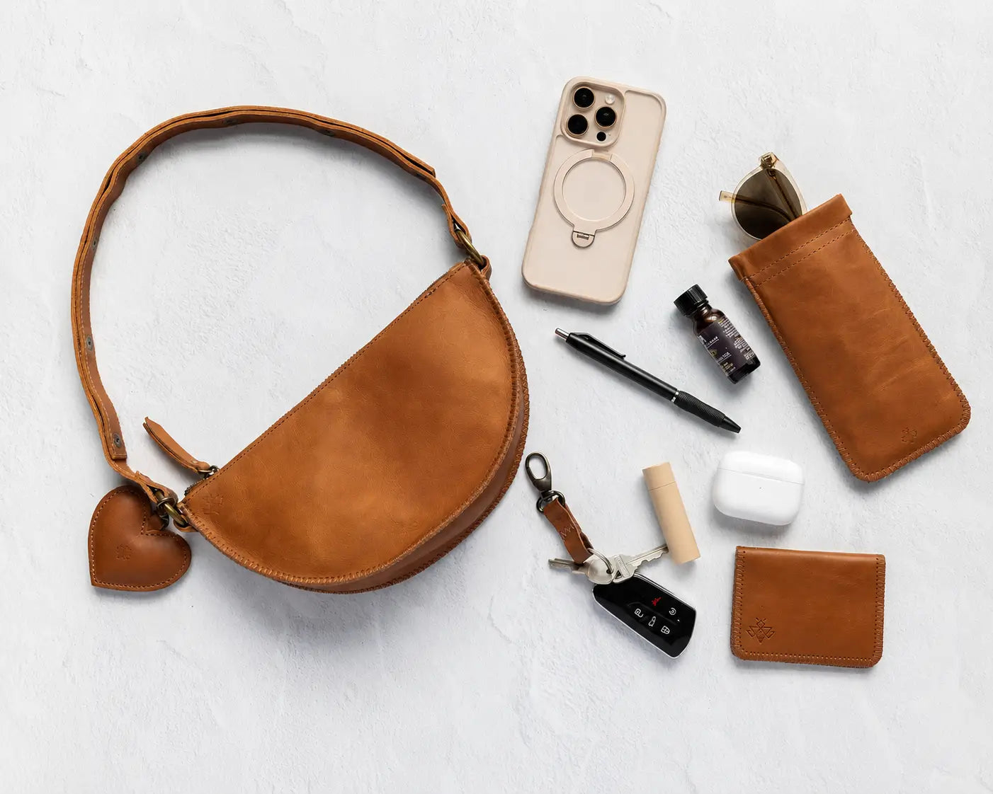 A warm tan leather half-moon shoulder bag with a heart-shaped charm, styled in a flat-lay arrangement with various accessories on a light gray textured background.