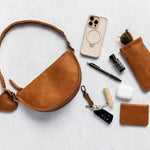 A warm tan leather half-moon shoulder bag with a heart-shaped charm, styled in a flat-lay arrangement with various accessories on a light gray textured background.