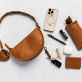 A warm tan leather half-moon shoulder bag with a heart-shaped charm, styled in a flat-lay arrangement with various accessories on a light gray textured background.