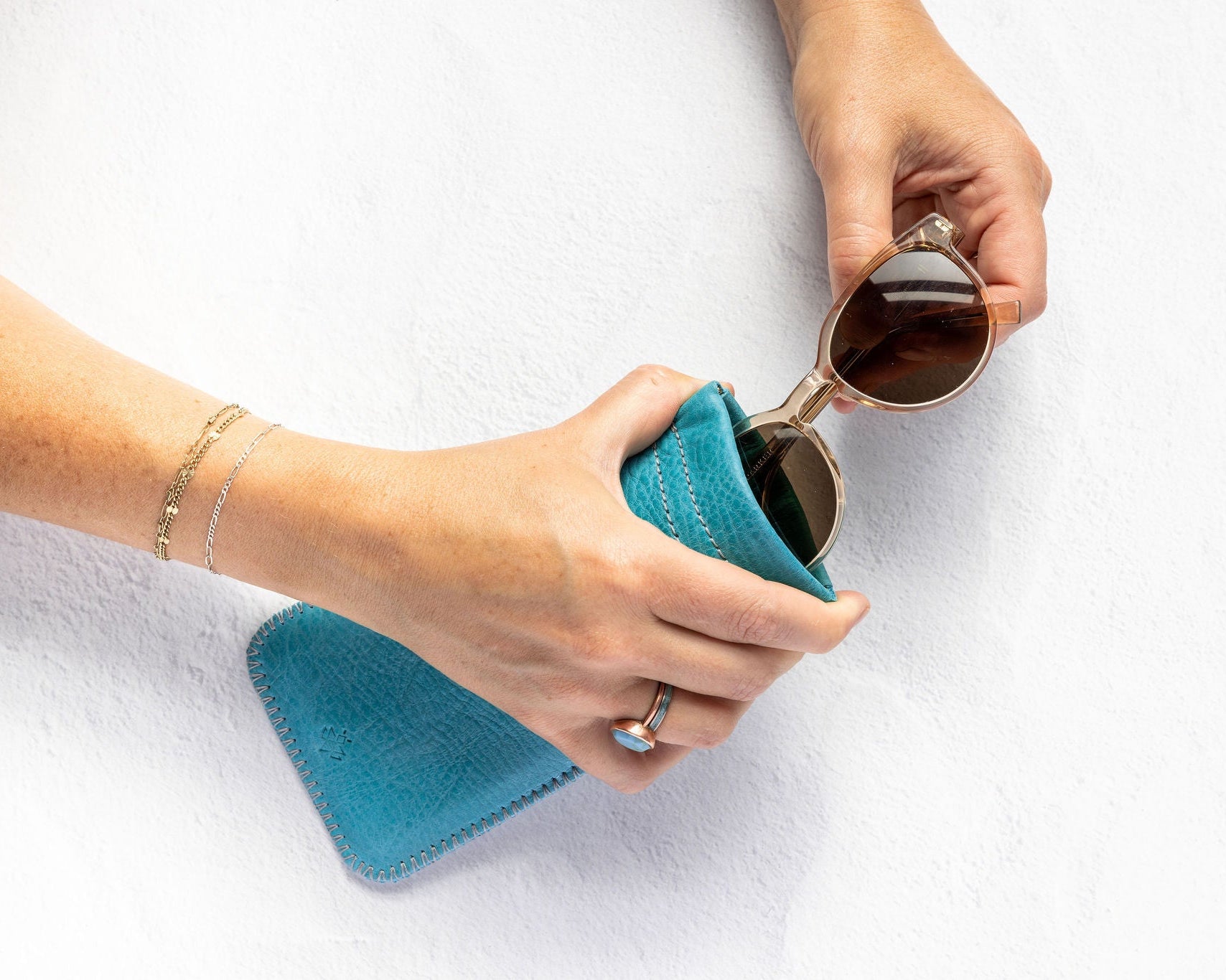 A person's hands holding a teal blue textured fabric pouch and placing round sunglasses into it, against a clean white textured background.