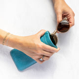 A person's hands holding a teal blue textured fabric pouch and placing round sunglasses into it, against a clean white textured background.