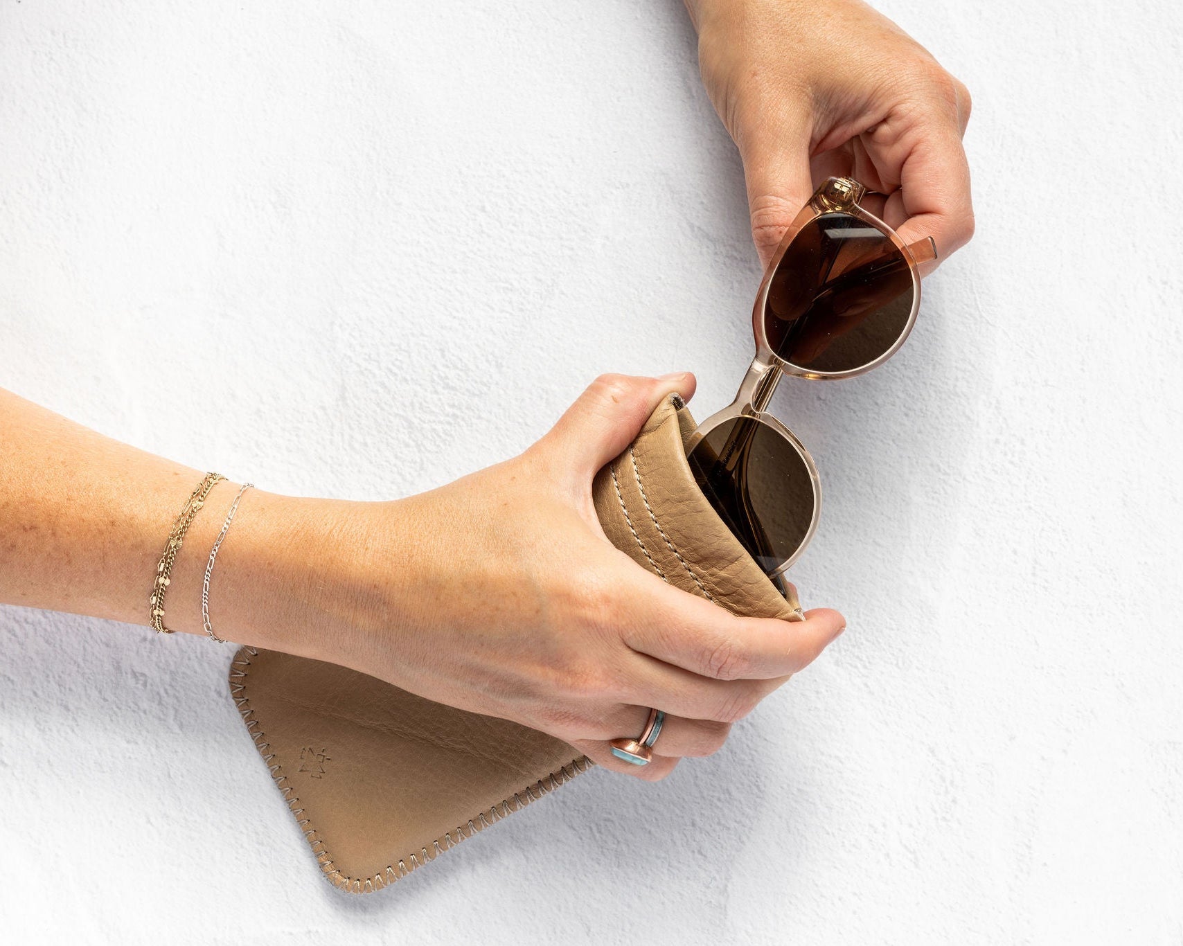 A person's hands are shown holding a pair of round sunglasses and placing them into a beige leather case against a white textured background.