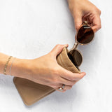 A person's hands are shown holding a pair of round sunglasses and placing them into a beige leather case against a white textured background.